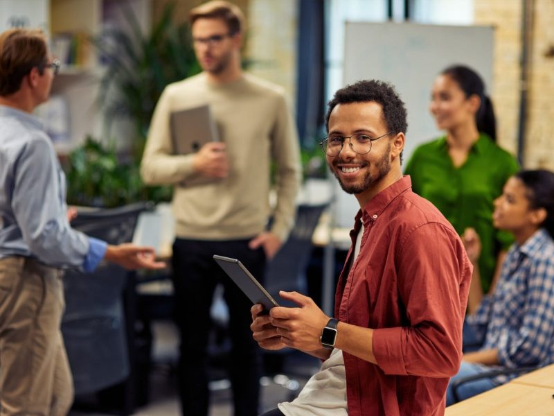 young-cheerful-mixed-race-man-holding-digital-tablet-and-smiling-at-camera-while-working-with.jpg